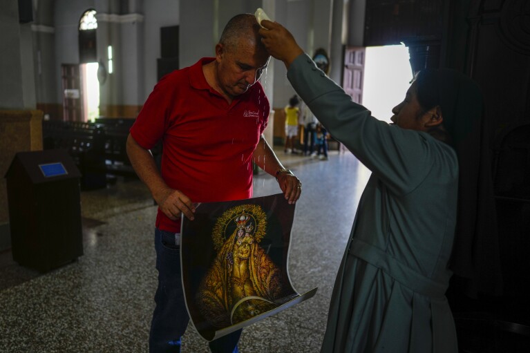 Ramon Nieblas is blessed by a nun with holy water as he holds a poster of the Virgin of Charity of Cobre at the shrine of the Cuban patron saint in El Cobre, Cuba, Sunday, Feb. 11, 2024. Nieblas, a Cuban living in Brazil who traveled thousands of miles to the basilica in eastern Cuba, asked the Virgin to save his son who has leukemia, and his wife who also has cancer. (AP Photo/Ramon Espinosa)