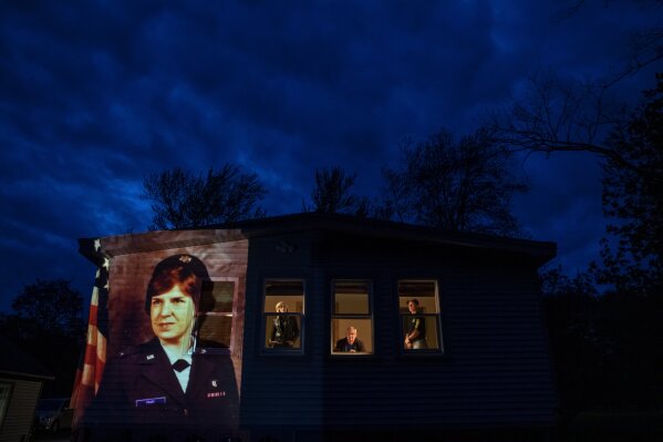 An image of veteran Constance "Kandy" Pinard is projected onto the home she grew up in with her sister, Tammy Petrowicz, left, and brothers, Paul, center, and Brian Driscoll in Florence, Mass., Thursday, May 14, 2020. Pinard, a nurse in the U.S. Air Force and resident of the Soldier's Home in Holyoke, Mass., died from the COVID-19 virus at the age of 73. Seeking to capture moments of private mourning at a time of global isolation, the photographer used a projector to cast large images of veterans on to the homes as their loved ones are struggling to honor them during a lockdown that has sidelined many funeral traditions. (AP Photo/David Goldman)
