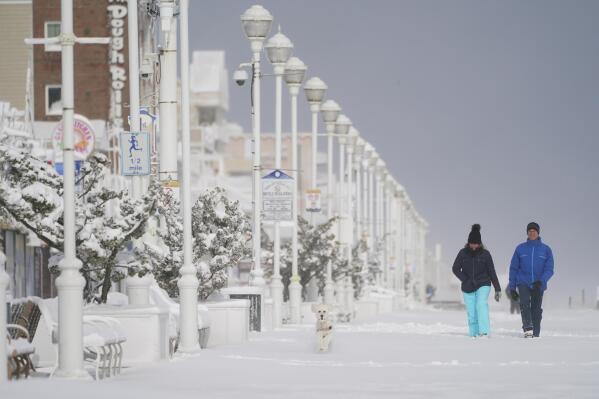 A dog is followed by a couple as the stroll on the snow-covered boardwalk, Saturday, Jan. 29, 2022, in Ocean City, Md. A powerful nor'easter swept up the East Coast on Saturday, threatening to bury parts of 10 states under deep, furiously falling snow accompanied by coastal flooding and high winds that could cut power and leave people shivering in the cold weather expected to follow. (AP Photo/Julio Cortez)