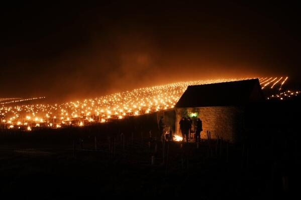 Varios viticultores se calientan en torno a una hoguera mientras velas colocadas para proteger los brotes y flores de la escarcha brillan en un viñedo en Chabli, Borgoña, el lunes 4 de abril de 2022. (AP Foto/Thibault Camus)