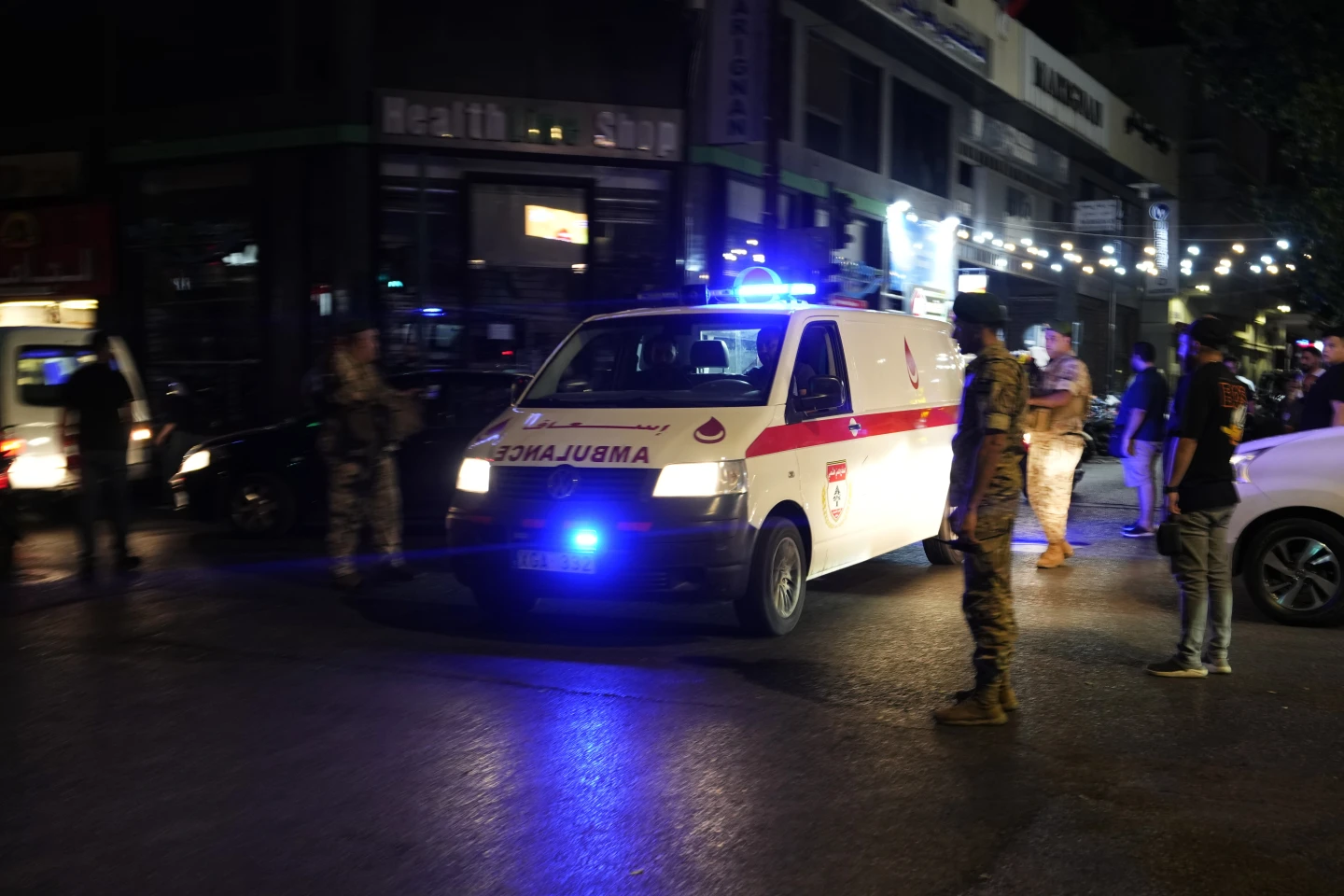 Una ambulancia blanca con franjas rojas, iluminada por luces de emergencia azules, estacionada en una calle de noche. Alrededor de la ambulancia, se ven varias personas, algunas en uniforme militar, vigilando la escena. El ambiente sugiere una situación de emergencia, con vehículos y luces brillantes en el fondo, posiblemente en una zona urbana.