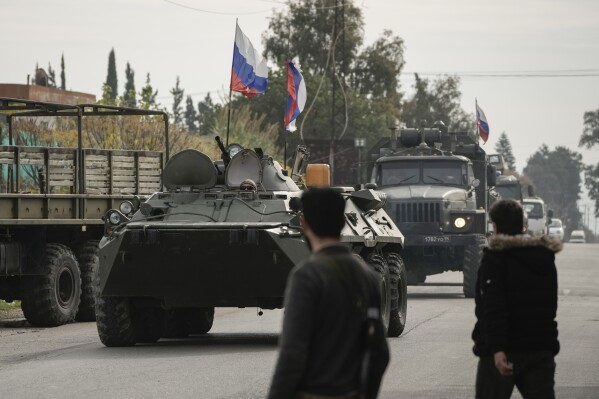 Syrian fighters watch Russian armoured vehicles driving past near the Hmeimim Air Base, a Syrian airbase currently operated by Russia, in the town of Hmeimim, southeast of Latakia, Syria, Monday Dec. 16, 2024.(AP Photo/Leo Correa, File)