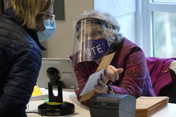 FILE - In this Oct. 28, 2020, file photo, poll worker Alice Machinist, of Newton, Mass., right, wears a mask and shield out of concern for the coronavirus while assisting a voter, left, with a ballot during early in-person general election voting at the Newton Free Library, in Newton, Mass. Masks are required at some polling places around the country and strongly encouraged in most others as a basic precaution to help keep poll works and others safe from the fast-spreading coronavirus. But mandates tying a face covering to casting a ballot are sure to lead to confrontations on Election Day, and those will almost certainly grab wide attention if they arise in any of the presidential battleground states. (AP Photo/Steven Senne, File)
