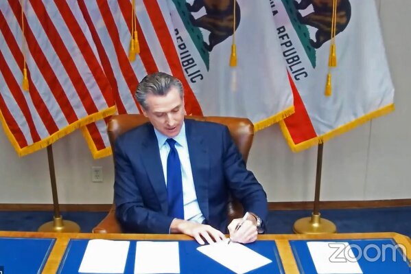 In this image taken during a video broadcast via Zoom, California Gov. Gavin Newsom signs a law on Friday, Sept. 25, 2020, in Sacramento, Calif.,  that for the first time defines “medical necessity," a move aimed at requiring private health insurance plans to pay for more mental health and drug addiction treatments.  (Zoom via AP)