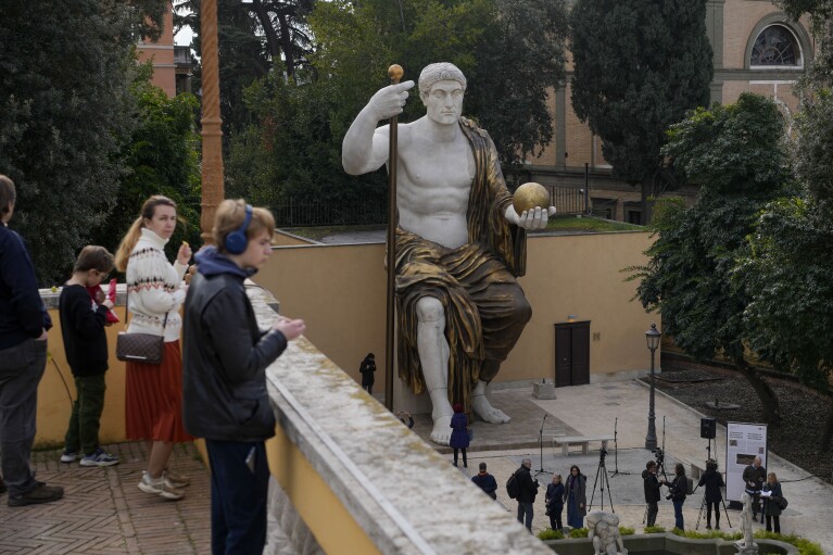 Visitors admire a massive, 13-meter (yard) replica of the statue Roman Emperor Constantine commissioned for himself after 312 AD that was built using 3D technology from scans of the nine giant original marble body parts that remain, as it was unveiled in Rome, Tuesday, Feb. 6, 2024. The imposing figure of a seated emperor, draped in a gilded tunic and holding a scepter and orb, gazing out over his Rome, is located in a side garden of the Capitoline Museums, just around the corner from the courtyard where the original fragments of Constantine's giant feet, hands and head are prime tourist attractions. (AP Photo/Andrew Medichini)