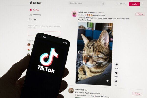 FILE - The TikTok logo is seen on a mobile phone in front of a computer screen which displays the TikTok home screen, Saturday, March 18, 2023, in Boston. European Union regulators said Wednesday, April 17, 2024, they're seeking details from TikTok on a new app from the video sharing platform that pays users to watch videos. (Ǻ Photo/Michael Dwyer, File)