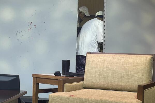 Blood stains mark a screen as author Salman Rushdie, behind screen, is tended to after he was attacked during a lecture, Friday, Aug. 12, 2022, at the Chautauqua Institution in Chautauqua, N.Y., about 75 miles (120 km) south of Buffalo. (AP Photo/Joshua Goodman)