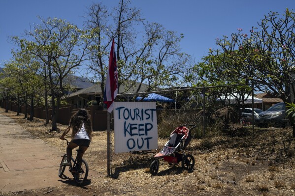 A girl rides her bike past a sign that says "Tourist Keep Out," in Lahaina, Hawaii, Thursday, Aug. 17, 2023. Long before a wildfire blasted through the island of Maui the week before, there was tension between Hawaii's longtime residents and the visitors some islanders resent for turning their beaches, mountains and communities into playgrounds. But that tension is building in the aftermath of the deadliest U.S. wildfire in more than a century. (AP Photo/Jae C. Hong)