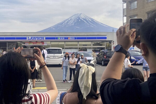Odwiedzający robią zdjęcie przed sklepem spożywczym w mieście Fujikawaguchiko w prefekturze Yamanashi w Japonii, na tle pana Fuji, 28 kwietnia 2024 r. Miasto Fujikawaguchiko, znane z wielu popularnych miejsc fotograficznych japońskiej marki Mt.  Fuji we wtorek, 30 kwietnia 2024 r., umieściła ogromny czarny ekran wzdłuż chodnika, aby zasłonić widok na góry w dzielnicy dotkniętej najnowszym przypadkiem nadmiernej turystyki w Japonii.  (Wiadomości Kyodo za pośrednictwem Associated Press)