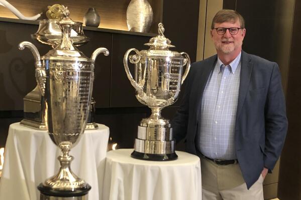 In this photo provided by Grace Vine, Beau Welling stands with PGA of America's championship trophies on Oct. 18, 2019, ahead of a preview event for the new PGA facility in Frisco, Texas. Welling is the golf course architect responsible for PGA Frisco's Fields Ranch West course. Like many late-to-the-game Americans, Beau Welling got hooked on the sport of curling after seeing the shouting and sweeping and clattering stones while watching the Olympics on television. (Beau Welling Design photo via AP)