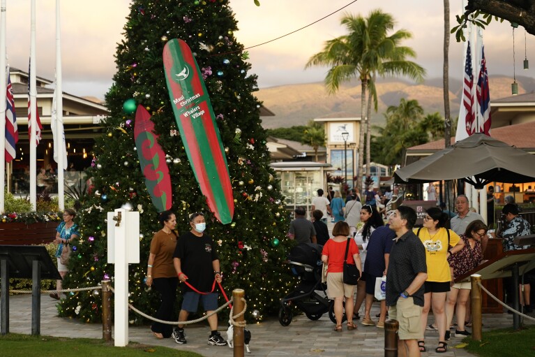 Visitors walk by a Christmas tree at Whalers Village, Wednesday, Dec. 6, 2023, in Lahaina, Hawaii. Residents and survivors still dealing with the aftermath of the August wildfires in Lahaina have mixed feelings as tourists begin to return to the west side of Maui. (AP Photo/Lindsey Wasson)