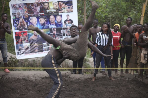 CORRECTS THE YEAR TO 2024 - Ugandan youths perform an amateur wrestling tangle in the soft mud in Kampala, Uganda Wednesday, March 20, 2024. The open-air training sessions, complete with an announcer and a referee, imitate the pro wrestling contests the youth regularly see on television. While a pair tangles inside the ring, made with bamboo poles strung with sisal rope, others standing ringside cheer feints and muscular shows of strength. (AP Photo/Patrick Onen)