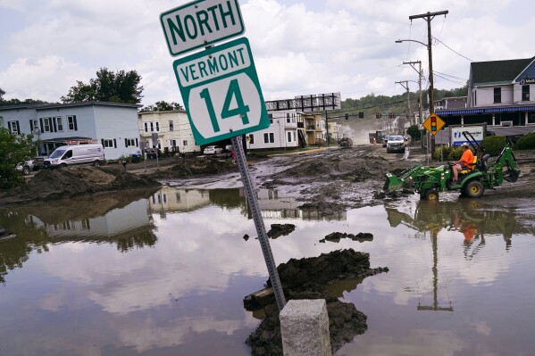 FILE - A small tractor clears water from a business as flood waters block a street, July 12, 2023, in Barre, Vt. Vermont has become the first state to enact a law requiring fossil fuel companies to pay a share of the damage caused by climate change, Thursday, May 30, 2024, after the state suffered catastrophic summer flooding and damage from other extreme weather. (AP Photo/Charles Krupa, file)