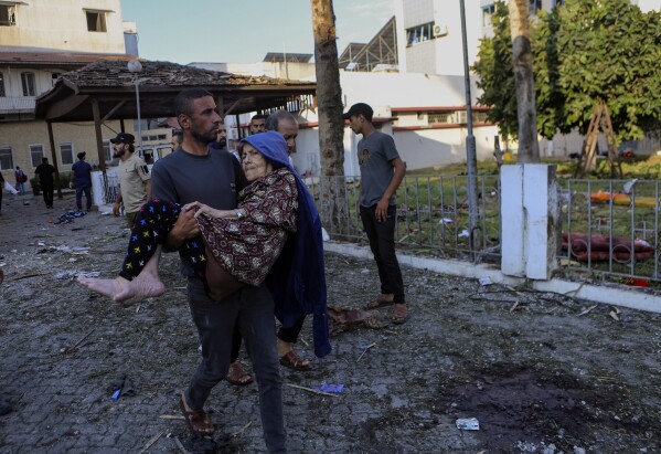 A Palestinian man carries an elderly woman past the site of a deadly explosion at al-Ahli hospital, in Gaza City, Wednesday, Oct. 18, 2023. (AP Photo/Abed Khaled)