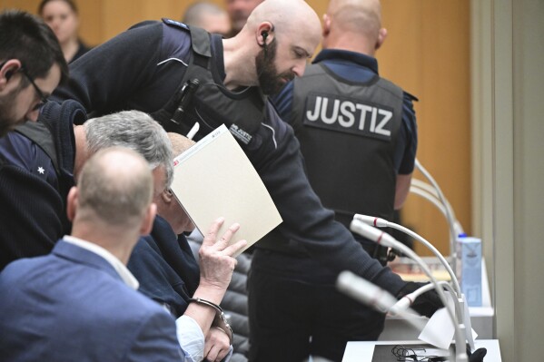 A defendant is led into the courtroom, at the Higher Regional Court at the start of a trial, in Stuttgart, Germany, Monday, April 29, 2024. Nine people charged in connection with an alleged far-right plot to topple the German government are going on trial in one of three cases linked to the plot that came to light in late 2022. (Bernd Wei'brod/dpa via AP)