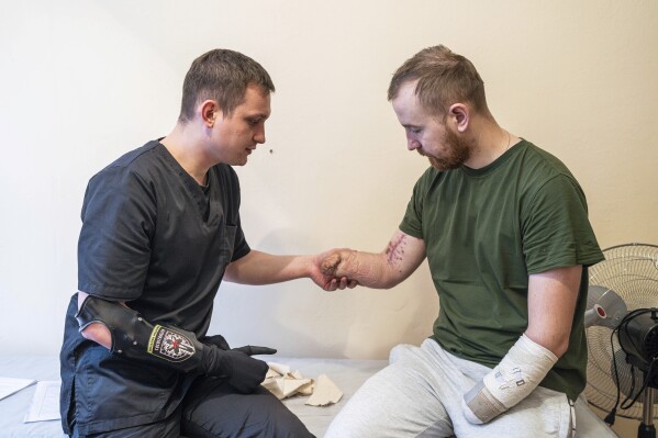 Dr. Alexis Cholas, left, examines the amputated limbs of Ukrainian soldier Volodymyr Symyshyn at the hospital in Kyiv, Ukraine, Wednesday, Nov. 8, 2023. Twenty-six-year-old Cholas lost his arm when, as part of a team of combat medics, he was evacuating the wounded, and the vehicle was involved in a traffic accident in eastern part of the country. This happened about a year ago, and four months after the injury, he received a bionic arm from Esper Bionics. (AP Photo/Evgeniy Maloletka)