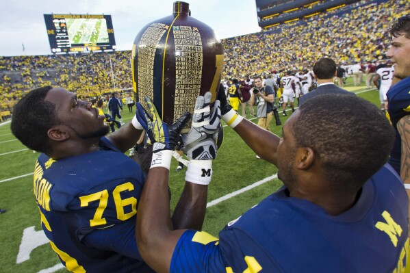FILE - Michigan defensive tackle Quinton Washington (76) and cornerback Courtney Avery (11) celebrate with the Little Brown Jug trophy after the team's win over Minnesota in an NCAA college football game Oct. 5, 2013, in Ann Arbor, Mich. Though Michigan's dominance in the series with Minnesota has stretched into a sixth decade and both teams have two other rivalries that are much fiercer, the jug is another piece of the rich history of the sport being the pushed to the background by the broadcast-driven expansion and realignment of the major conferences. (AP Photo/Tony Ding, File)