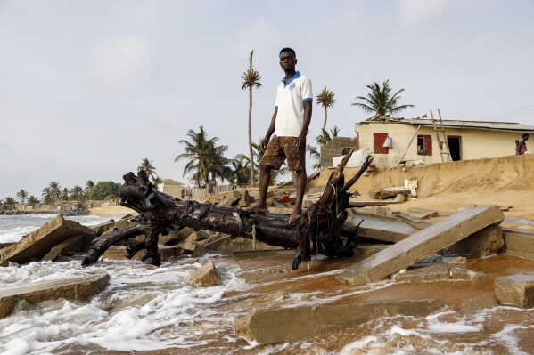 Dennis Dostey Dorve, a driver and a part-time fisherman, stands on a fallen tree next to his late father's room, collapsed by the sea in the family home in Avegadzi, Ghana, Wednesday, March 5, 2025 (AP Photo/Misper Apawu)