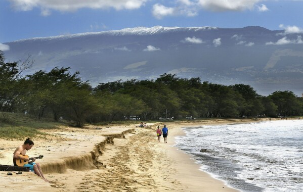 FILE -- Snow-capped Haleakala serves as a backdrop as Paia, Maui's Scott Picton, left, plays a guitar and Saskatchewan's Shannon and Dan Runcie walk on Sugar Beach in South Maui, Hawaii, Feb. 11, 2019. Local officials on the Hawaiian island of Maui on Wednesday, June 5, 2024, voted to oppose a U.S. military proposal to build new telescopes on the summit of Haleakala volcano. (Matthew Thayer/The Maui News via AP)