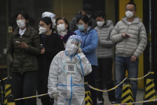 A worker in a protective suit talks on her phone as residents wearing face masks stand in line for their routine COVID-19 tests at a testing site in Beijing, Thursday, Nov. 24, 2022. (AP Photo/Andy Wong)