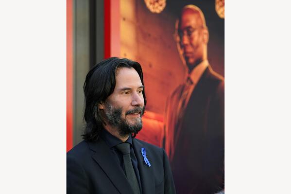 Keanu Reeves, the star of "John Wick: Chapter 4," wears a blue ribbon on his lapel in tribute to his late fellow cast member Lance Reddick, seen in a poster in the background, at the premiere of the film, Monday, March 20, 2023, at the TCL Chinese Theatre in Los Angeles. The veteran character actor Reddick died Friday morning at age 60. (AP Photo/Chris Pizzello)