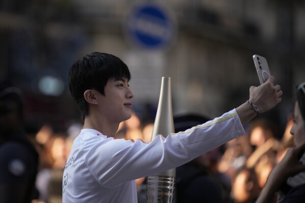 Jin, the oldest member of K-pop supergroup BTS, males a selfie with the Olympic torch, Sunday, July 14, 2024 in Paris. Jin will carry the Olympic torch in the area of Paris' Louvre Museum after recently completing his mandatory military service. (AP Photo/Louise Delmotte)
