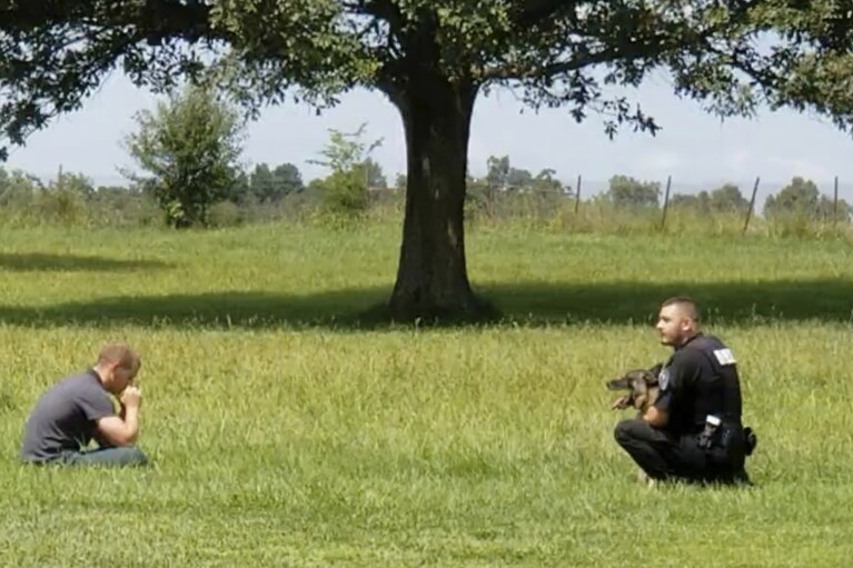 In this image from video provided by a family friend, Taylor Ware, left, sits in a field approached by a police officer and canine at a highway rest stop in Dale, Ind., on Aug. 25, 2019. Taylor's mother called 911 when he wouldn’t get back in their SUV during a manic episode caused by bipolar disorder. (Pauline Engel via AP)