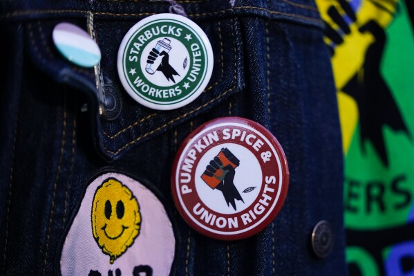 FILE - Employee Quint Palermo wears union pins on their jacket as Starbucks workers participate in a walkout and strike during the company's Red Cup Day Thursday, Nov. 16, 2023, at the company's first Reserve roastery in Seattle. The federal government is delaying a new rule that could make it easier for millions of workers to unionize after business groups sued. The National Labor Relations Board says the rule, which was scheduled to go into effect next month, will now be effective Feb. 26 to give it time to resolve legal challenges. (AP Photo/Lindsey Wasson, File)