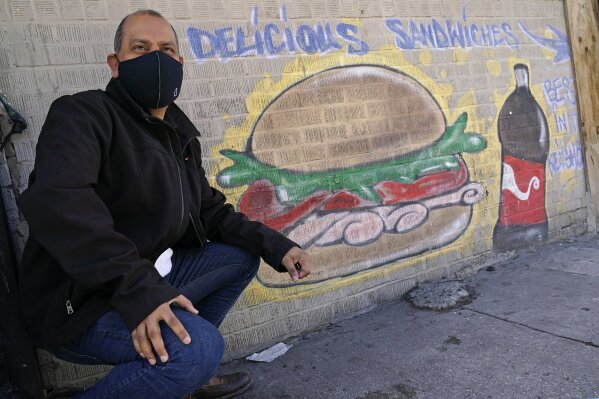Bodega owner Francisco Marte poses in front of a sign outside his deli-style store, Wednesday, Feb. 10, 2021, in the Bronx borough of New York. Marte president of the Bodega and Small Business Association of New York, which represents bodegas in New York, says he's lobbied local officials to set aside COVID-19 vaccine appointments for bodega workers, many of whom are unaware they are eligible.(AP Photo/Kathy Willens)