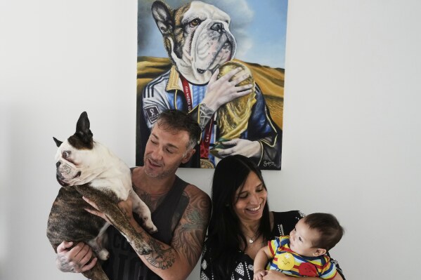 Carolina Morales holds her four-month-old son Benjamin as her husband Alejandro Tirachini holds the family pet Thay, whom they consider their first son, while they pose for a photo backdropped by a painting of Thay by artist Lisandro Guma, in Buenos Aires, Argentina, Friday, April 11, 2025. (AP Photo/Natacha Pisarenko)