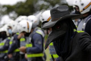 A demonstrator dressed in a bird mask, representing the black plague, stands near police guarding a city government office where demonstrators protest a two-week-long lockdown to curb the spread of COVID-19 in Brasilia, Brazil, Monday, March 1, 2021. It’s the second lockdown in Brasilia since the start of the pandemic one year ago. (AP Photo/Eraldo Peres)
