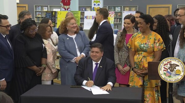 In this screenshot from a livestream broadcast by the State of Illinois, Gov. J.B. Pritzker signs a bill, Monday, June 12, 2023, at Harold Washington Library's Thomas Hughes Children's Library in downtown Chicago. The new law will require the state's libraries to uphold a pledge not to ban material because of partisan disapproval, starting on Jan. 1, 2024. If they refuse, they will not receive state funding. Pritzker said the law will make Illinois the first state in the nation to outlaw book bans. (State of Illinois via AP)