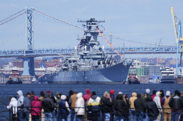 People gather in Gloucester City, N.J., to view the USS New Jersey moved down Delaware River Thursday, March 21, 2024. The retired battleship is initially headed to the Paulsboro Marine Terminal, where it will be balanced to prepare for dry docking, and will then go to the Philadelphia Navy Yard. (AP Photo/Matt Rourke)