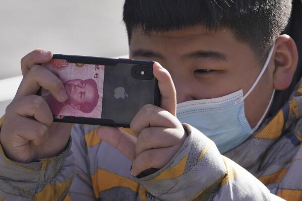 A boy wearing a face mask using an iPhone with a Chinese currency note takes a picture on a street in Beijing, Tuesday, Jan. 17, 2023. China's economic growth fell to its second-lowest level in at least four decades last year under pressure from anti-virus controls and a real estate slump, but activity is reviving after restrictions that kept millions of people at home and sparked protests were lifted. (AP Photo/Andy Wong)