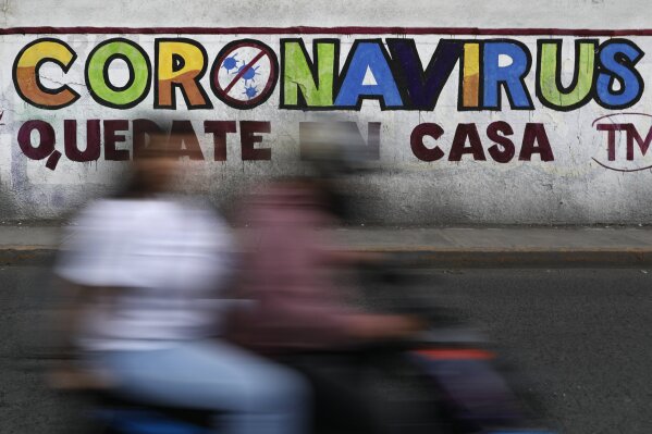 In this April 28, 2020 photo, a couple rides a motorcycle past a painted sign reading in Spanish "Coronavirus. Stay Home," in Iztapalapa, Mexico City. Mexico has had extremely limited testing compared with other countries, another factor that experts have said made it difficult for the country to contain the virus. (AP Photo/Rebecca Blackwell)