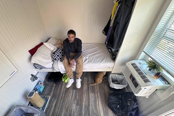 Eric Martinez sits in his room at a micro community in Denver, Wednesday, June 5, 2024. Martinez along with the others were directed into the micro communities of small cabin-like structures with a twin bed, desk and closet, after his Denver tent encampment was swept. The city built three such communities with nearly 160 units total in about six months, at roughly $25,000 per unit. (AP Photo/Thomas Peipert)