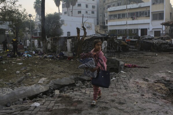 A Palestinian girl carries a blankets as she walks past the site of a deadly explosion at al-Ahli hospital, in Gaza City, Wednesday, Oct. 18, 2023. (AP Photo/Abed Khaled)