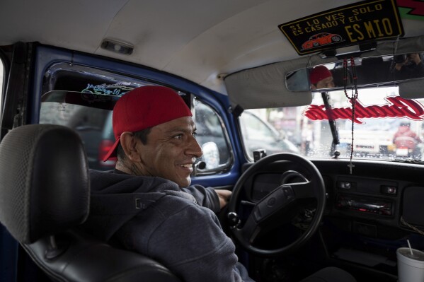 Taxi driver Claudio Garcia sits inside his Volkswagen Beetle that he has named "Gualupita" behind his wife, in the Cuautepec neighborhood of Mexico City, Friday, June 21, 2024. As some of the older Beetles rock together, the paint long faded from years of wear and tear, other drivers like Garcia dress up their cars, kept them inside top form.  (AP Photo/Aurea Del Rosario)