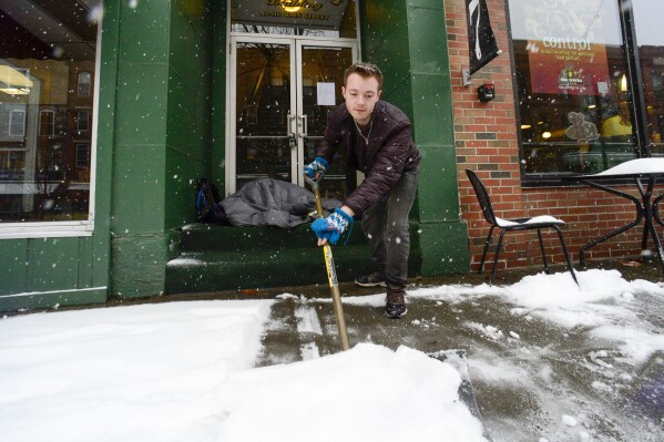 Mikey Reynolds, an employee at The Works on Main Street in Brattleboro, Vt., shovels the sidewalk in front of the restaurant as a person who is facing homelessness sleeps in a door entryway while seeking refuge from the snow storm on Saturday, March 23, 2024. New England is battling a mix of wind, rain, sleet and heavy snow across the region Saturday with more than a foot of snow expected in ski county, but mostly rain, wind and possible flooding in southern areas and along the coast. (Kristopher Radder /The Brattleboro Reformer via AP)