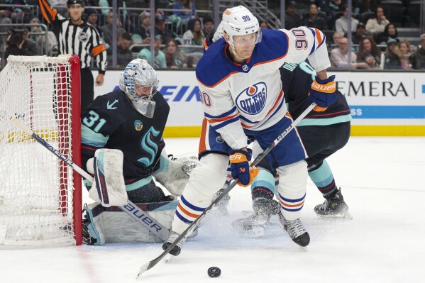 Edmonton Oilers right wing Corey Perry (90) moves the puck as Seattle Kraken goaltender Philipp Grubauer (31) and defenseman Brian Dumoulin (8) defend during the second period of an NHL hockey game Saturday, March 2, 2024, in Seattle. (AP Photo/Jason Redmond)