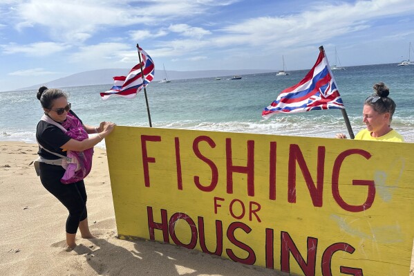 Jordan Ruidas, left, and Katie Austin adjust a sign on Kaanapali Beach in Lahaina, Hawaii on Wednesday, Nov.14, 2023. A group of Lahaina wildfire survivors is vowing to camp on a popular resort beach until the mayor uses his emergency powers to shut down unpermitted vacation rentals and make the properties available for residents in desperate need of housing. (AP Photo/Audrey McAvoy)