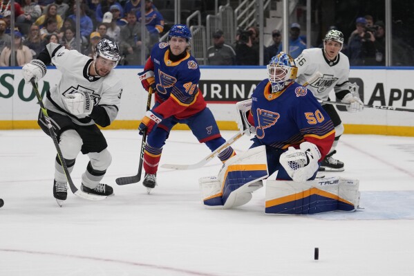 St. Louis Blues goaltender Jordan Binnington (50) deflects a puck as teammate Scott Perunovich (48) and Los Angeles Kings' Adrian Kempe (9) watch during the first period of an NHL hockey game Wednesday, March 13, 2024, in St. Louis. (AP Photo/Jeff Roberson)