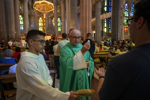 A priest holds a holy host while giving communion to faithful during a Mass in the Sagrada Familia basilica in Barcelona, Spain, Sunday, July 9, 2023. With tourism reaching or surpassing pre-pandemic levels across Southern Europe this summer, iconic sacred sites struggle to find ways to accommodate both the faithful who come to pray and millions of increasingly secular visitors attracted by art and architecture. (AP Photo/Emilio Morenatti)