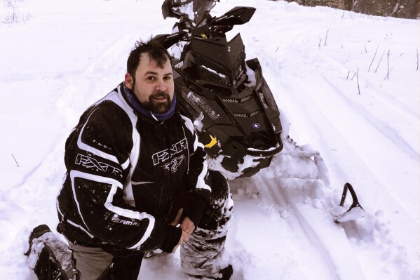 In this photograph provided by Richard Smith, Jeffrey Smith poses with a snowmobile during March 2018, in Windsor, Mass. Jeffrey Smith has filed suit against the government to pay nearly $10 million after being badly injured in a snowmobile crash in 2019 with a Black Hawk helicopter. Smith's snowmobile collided with a helicopter that was parked on a Massachusetts snow-covered trail at dusk. (Jeffrey Smith photo via AP)