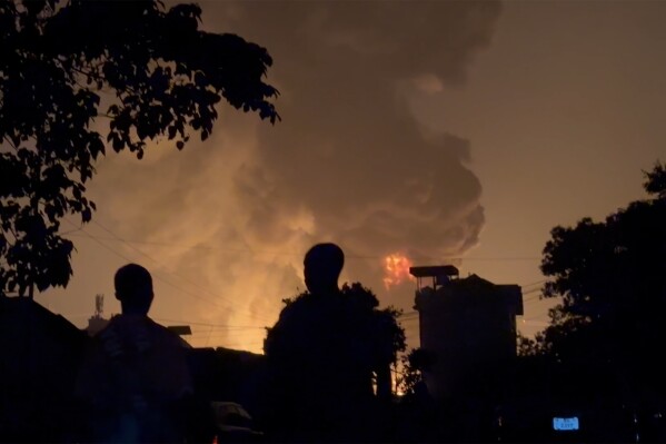 In this image made from video, residents watch a plume of smoke from a burning oil depot, in Conakry, Guinea, Monday, Dec. 18, 2023. An explosion and inferno at Guinea’s main fuel depot in the capital of Conakry has left several people dead or injured. Guinea’s presidency says the fire broke out at the Guinean Petroleum Company depot shortly after a massive explosion past midnight Sunday. (AP Photo)