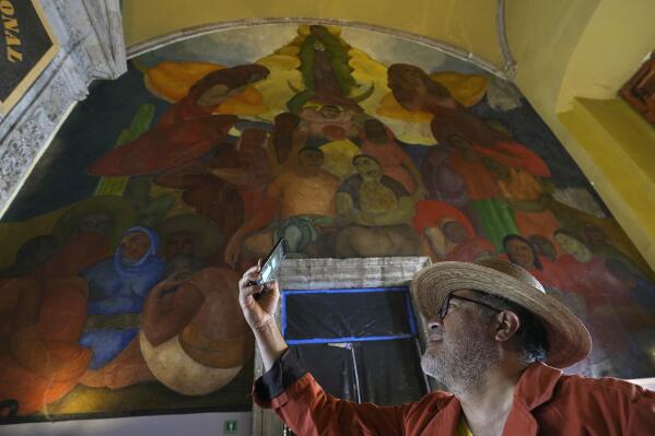 A tourist takes a photo backdropped by the "Alegoria de la Virgen de Guadalupe" mural, in the main entrance of the Antiguo Colegio de San Ildefonso, in Mexico City, Wednesday, April 26, 2023. The mural was created by Mexican artist Fermin Revueltas between 1922 and 1923, when the walls of San Ildefonso became the canvases where the muralist movement came to life. (AP Photo/Marco Ugarte)