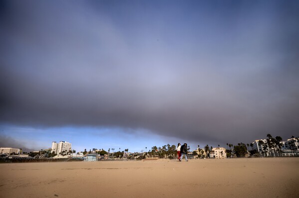 A couple walk along Santa Monica beach as a heavy plume of smoke from a wildfire burning in the Pacific Palisades passes over in Santa Monica, Calif. on Wednesday, Jan. 8, 2025. (AP Photo/Richard Vogel)