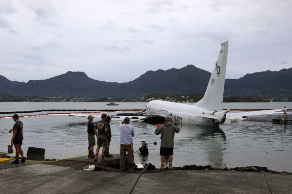 Contractors place inflatable bags under a U.S. Navy P-8A in Kaneohe Bay, Hawaii, Friday, Dec. 1, 2023, so they can float the aircraft over the water and onto land. The Navy plans to use inflatable cylinders to lift the jet off a coral reef and then roll it over to a runway to remove the plane from the ocean where it crashed the week before. (AP Photo/Audrey McAvoy)