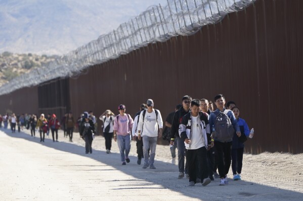 A group of people, including many from China, walk along the wall after crossing the border with Mexico to seek asylum, Tuesday, Oct. 24, 2023, near Jacumba, Calif. A major influx of Chinese migration to the United States on a relatively new and perilous route through Panama's Darién Gap jungle has become increasingly popular thanks to social media. (AP Photo/Gregory Bull)