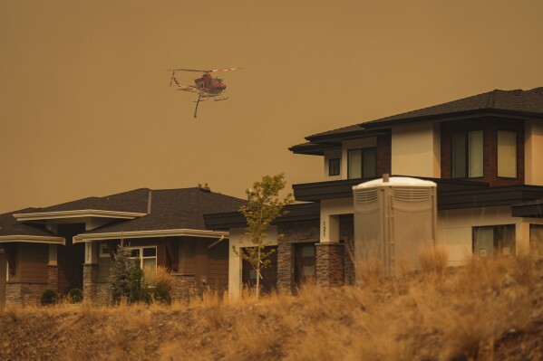 A helicopter equipped to respond to wildfires flies past homes in the Wilden neighbourhood near Knox Mountain after RCMP officers enforced a new evacuation order in Kelowna, British Columbia, Friday, Aug. 18, 2023. (Darryl Dyck/The Canadian Press via AP)